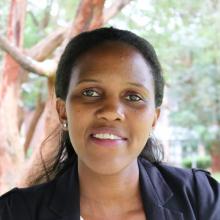 Headshot of Sarah Nyawira taken in natural light with trees in the background in the distance. Her black hair is pulled back in a half ponytail style and she smiles gently at the camera wearing a black blazer and diamond stud earrings.