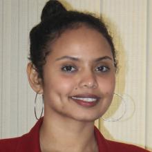 Headshot of Sam Said taken in front of a white textured backdrop. She smiles at the camera wearing a red blazer, large thin silver hoop earrings, and her dark hair is pulled up in a bun at the top of her head.