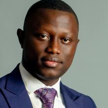 Headshot of Tamba Sangbah on a gray background. He looks at the camera with a slight smile. He wears a purple blazer, white collared shirt, and purple paisley tie. His hair is closely cropped and he is clean shaven.