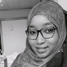 Headshot of Maimouna Seck in black and white. She wears a hajib and dark square-framed glasses, and smiles slightly at the camera.