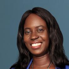 Headshot of Tendra Tenwah-Gweh against a dark blue-gray background. She smiles to the camera and wears her long dark hair straight. She wears a blue blouse and a silver chain.