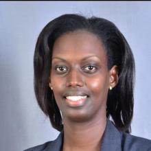Headshot of Sheila Uwase taken on a light blue backdrop. She smiles to the camera and wears a dark blue blazer, round gold earrings, and her dark straight hair is worn in a bob just above her shoulders.
