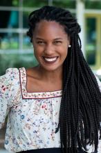 Andréia Coutinho Louback smiles broadly in this 2021 headshot. Her black hair is worn in long braids with some pulled back from her face. She wears large white pearl earrings, and a white square-neck blouse trimmed in red and decorated with blue and red flowers. 