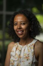 Ruth Tekle Andemariam smiles in a 2021 headshot. Her curly black hair is worn down and falls to nearly her shoulders. She wears small gold hoop earrings and a white blouse with a high pleated neck with orange and yellow flowers.