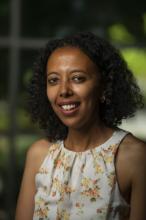 Ruth Tekle Andemariam smiles in a 2021 headshot. Her curly black hair is worn down and falls to nearly her shoulders. She wears small gold hoop earrings and a white blouse with a high pleated neck with orange and yellow flowers. 