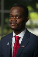 Sytekon Caspar Tarty smiles slightly in this 2021 headshot. His black hair is cropped close and he has a goatee. He wears a navy blazer, white collared shirt, red tie with a diamond patter, and a pin with the American and Liberian flags on his left lapel.