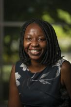 Sheila Apopo smiles broadly in this 2021 headshot. Her black hair is work in braids that reach her shoulders. She wears a sleeveless black blouse with large gray lilies. 