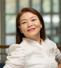 Samtso stands inside with the windows of the International Center behind her. She wears a white button-up blouse and gives a friendly smile to the camera.