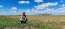 Patricia crouches in a grassy field in a white t shirt and jeans with blue skies and clouds in the background.