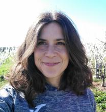 A smiling woman with wavy hair stands in an orchard, sunlight shining behind her.