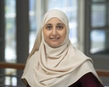 Headshot photo of a smiling woman in a beige hijab with blurred windows in the background.