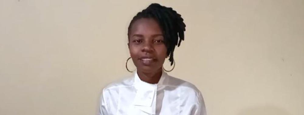 Esther Anyona stands before a white wall with her hands clasped in conversation. She smiles slightly and wears a white silk blouse with ruffles down the front, large gold hoop earrings, and her black hair is worn in locs pulled up to the top of her head and styled to her left.