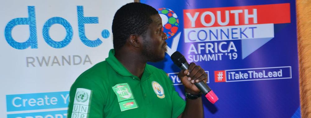 Jireh Bookwa stands before banners for Youth Connekt Africa Summit 2019. He holds a microphone as he speaks to the crowd, and wears a green polo shirt with various patches. His black hair is closely cropped, and he is in profile as he looks to his left.
