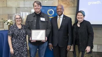 Four people smiling as a man in the center holds a framed certificate