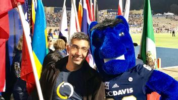 An international student holding an international flag smiles while standing next to Gunrock, UC Davis' blue mustang mascot