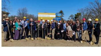 A group of people wearing hard hats and holding shovels participate in a groundbreaking