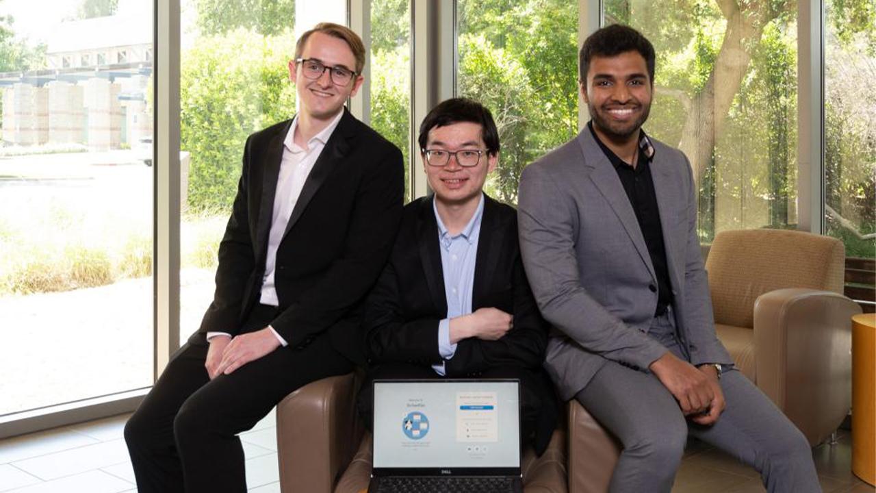 The winners sit on an arm chair inside a lobby with glass windows. An open laptop on Henry's lap displays the SchedGo website.