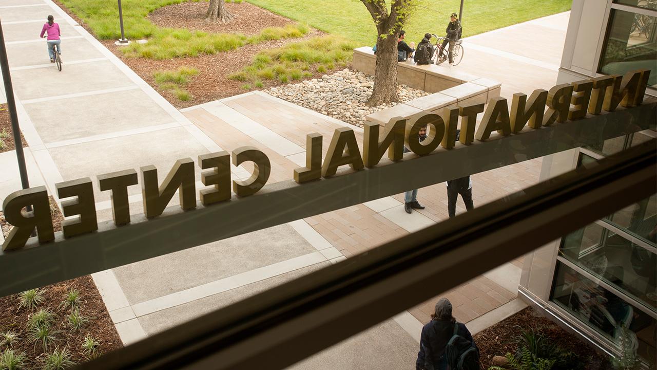 A photo from inside the International Center on the UC Davis campus looking out the main entrance window, taken on a fall day. The International Center sign is backwards from this vantage point looking out as a student wearing a hat, blue jacket and backpack walks away from the doors. Three other students, one sitting on a bike and two others sitting, chat under a tree outside. A student in a pink sweater rides her bike away from the building.