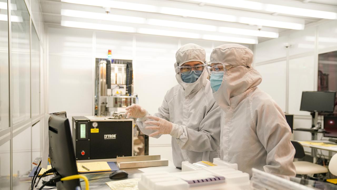 Two scientists in full cleanroom suits pointing at a computer monitor in a sterile lab.
