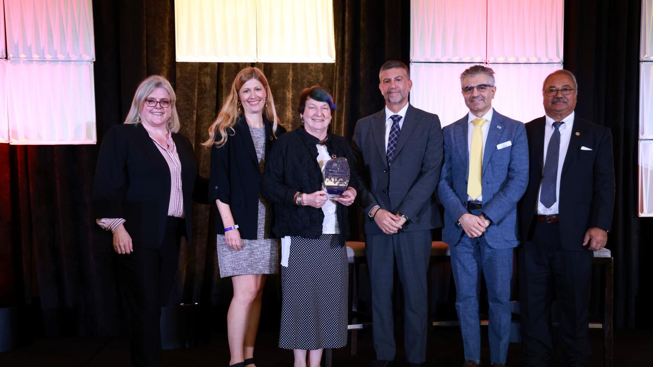 Six professionals stand on a stage, smiling. The woman in the center holds an award.