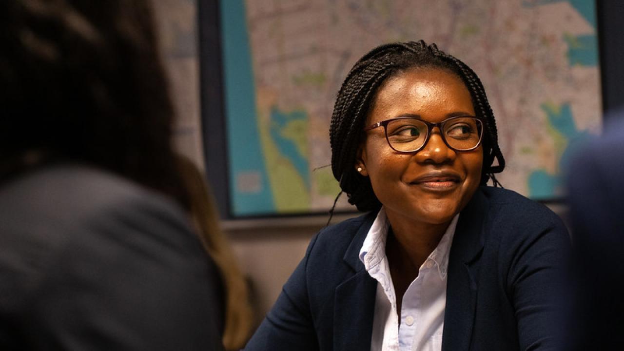 Lavinia Mbongo, a Mandela Washington Fellow, wears a white shirt and navy blazer with dark framed glasses. She is seated at a table, listening.