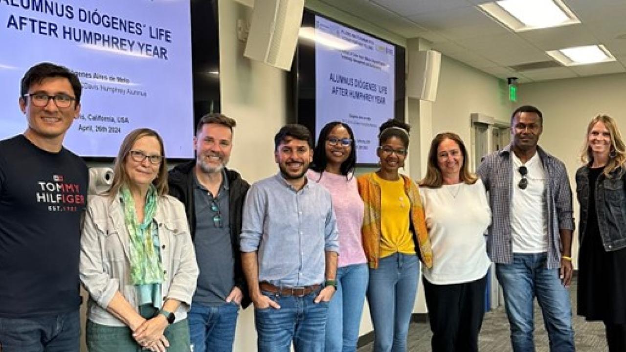 Nine people stand for a posed group picture in a room with two monitors that read "Alumnus Diogenes Life After Humphrey Year."