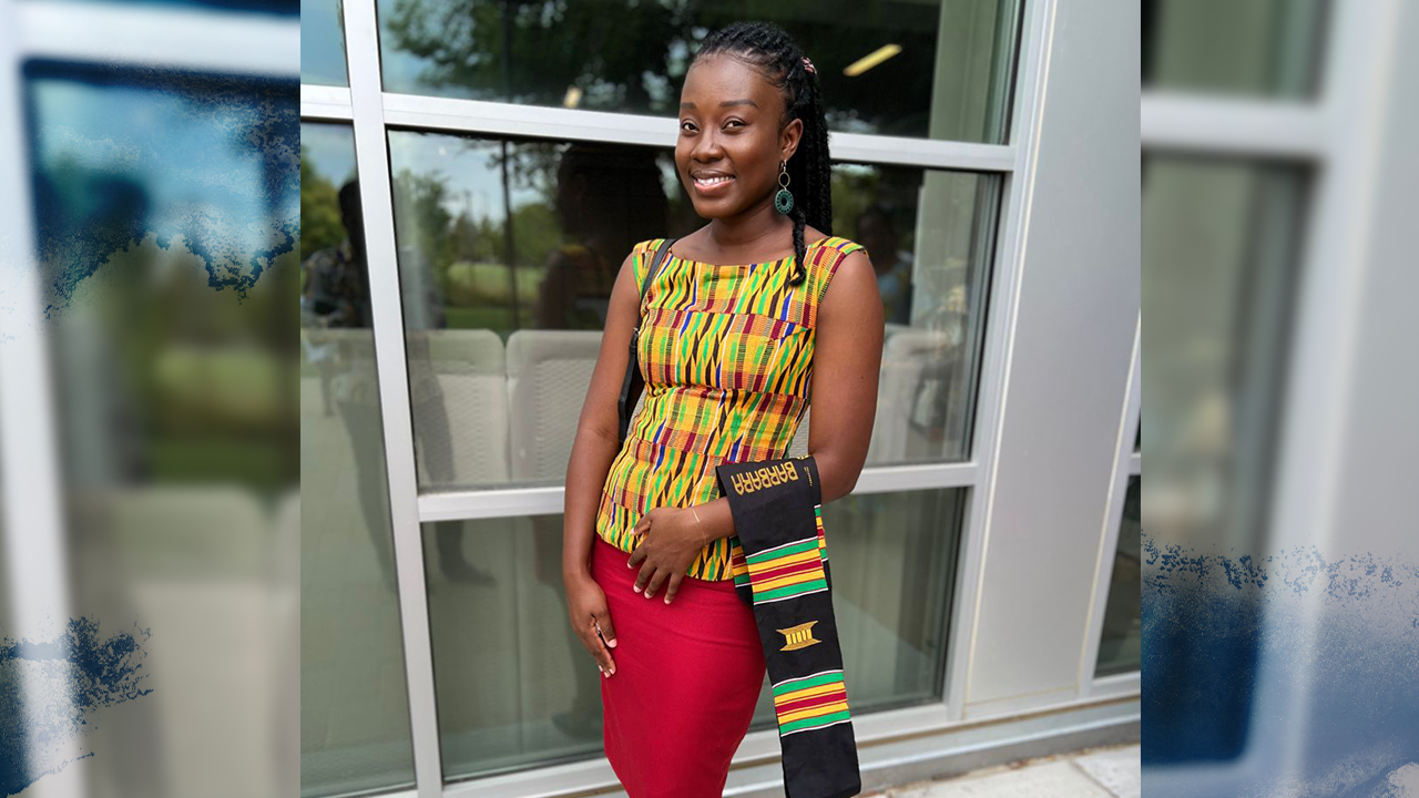Barbara Kumi stands outside in a shaded area in front of windows wearing a patterned sleeveless tunic in yellow, black, red, green and blue, and a red pencil skirt. Her hands are clasped in front of her and a black sash with the Ghana flag is draped over her left forearm.