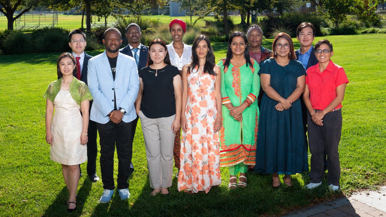 The 12 Humphrey Fellows stand posed in two rows outside on grass with an open lawn and bushes in the background.