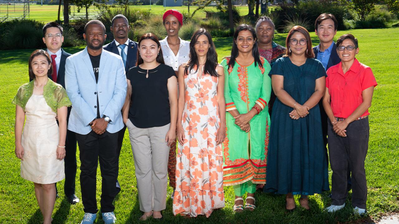 Twelve Humphrey Fellows stand together in two rows on the grass of a lawn on the UC Davis campus in the sunlight. They wear a combination of culturally traditional and business dress.
