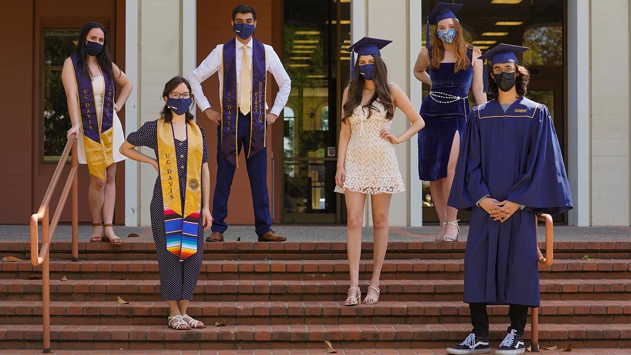 Six students wear commencement regalia and stand socially distanced wearing face masks on the steps of Mrak Hall on the UC Davis campus.