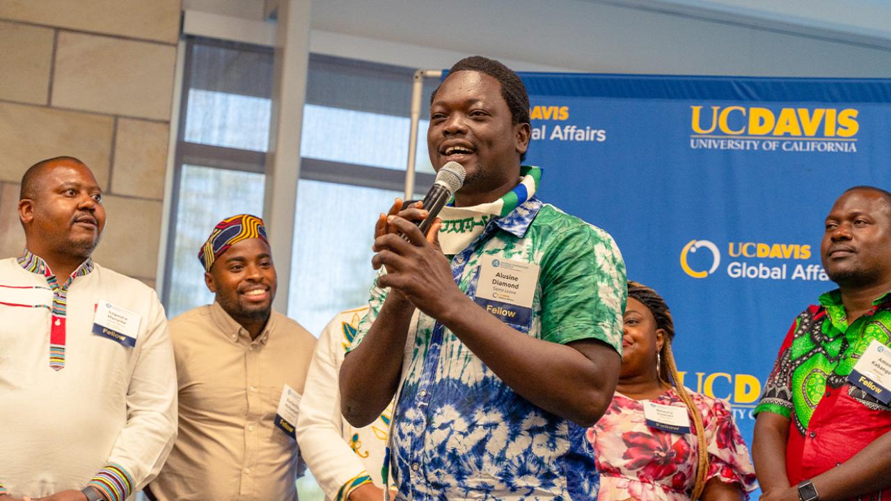 Diamond Suma stands in front of four Mandela Washington Fellows and speaks into a microphone. Behind them is a large sign with the UC Davis and UC Davis Global Affairs logos.
