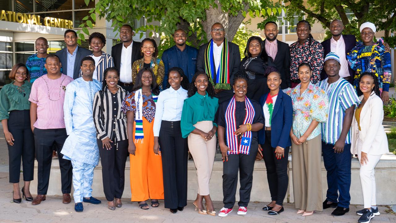 The 24 Fellows stand in two rows, with the back row elevated, under the shade of a large tree. Fellows wear a combination of business casual and cultural attire.