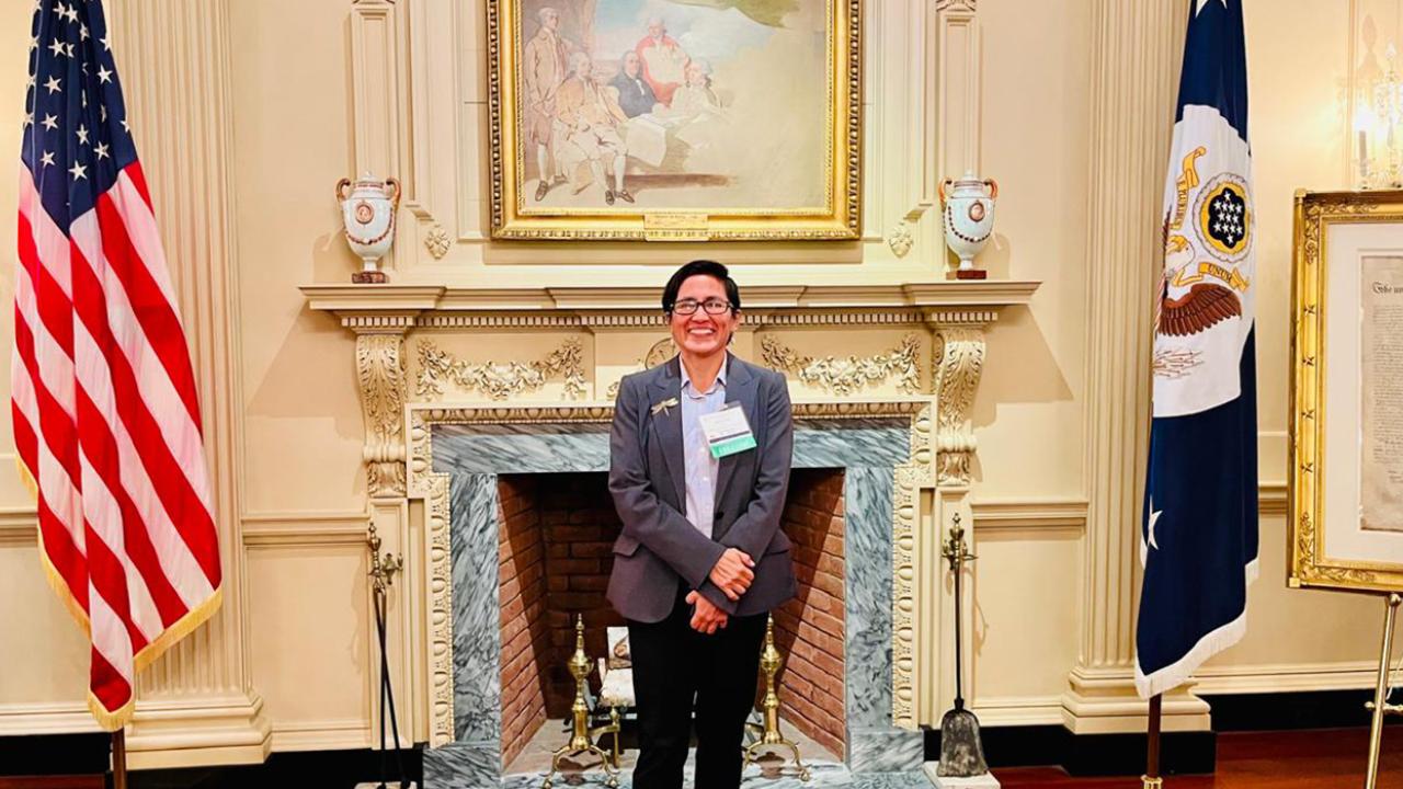 Ana stands in front of an ornate white fireplace and mantel. Over the fireplace is a classic painting and on either side of it are two flags: On the left is the American flag and on the right is the flag of the Office of the President of the United States of America.