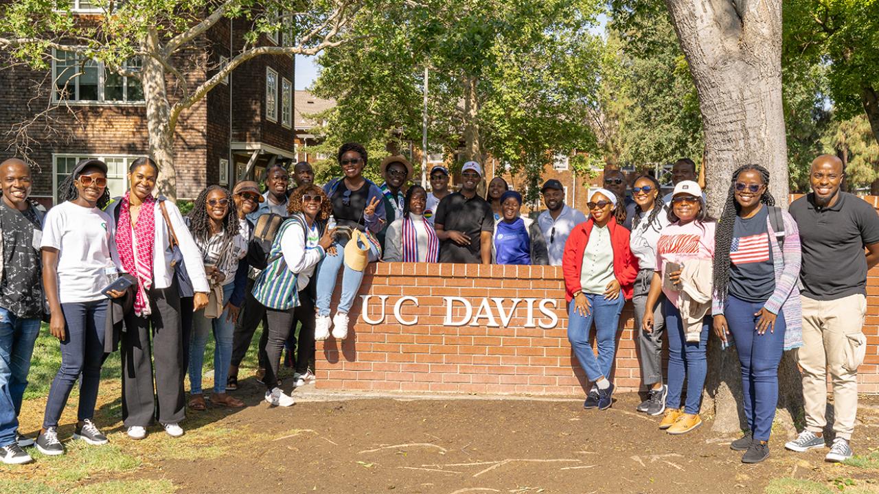 The cohort of 24 Mandela Washington Fellows gathers around a UC Davis sign on a short brick wall on the UC Davis campus.