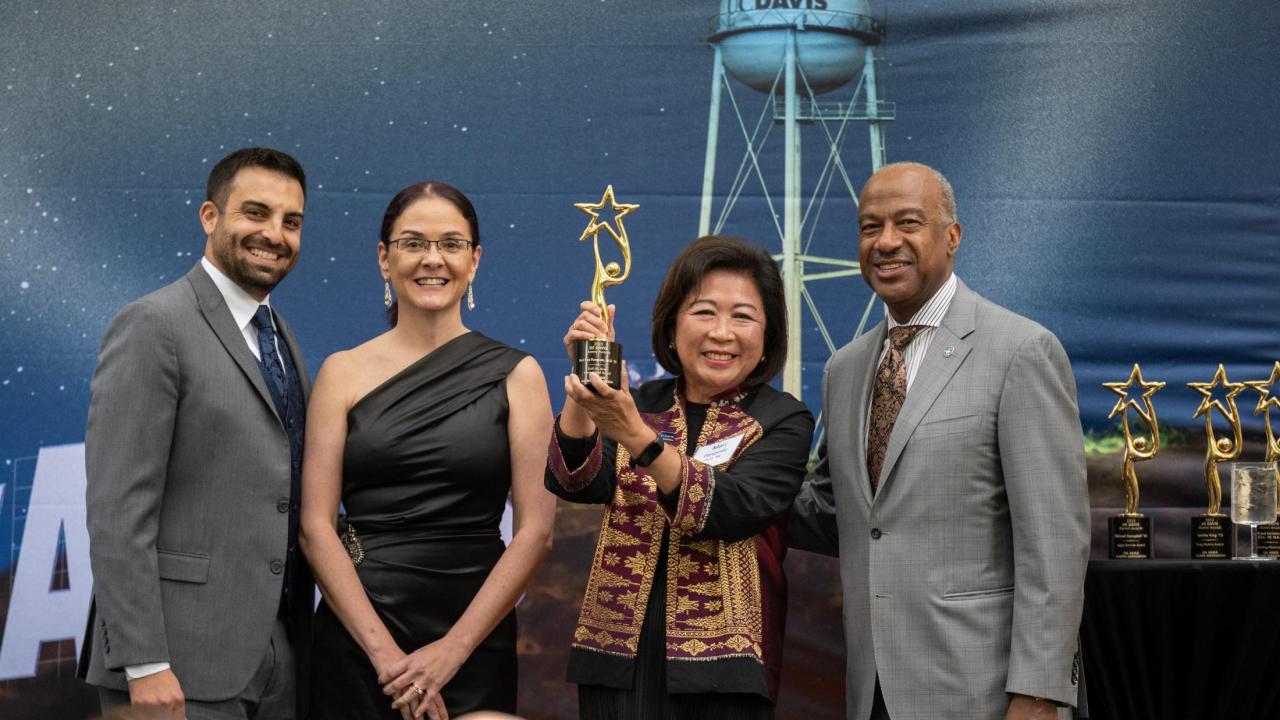 Four smiling people at awards ceremony, woman holding trophy, blue water-tower backdrop