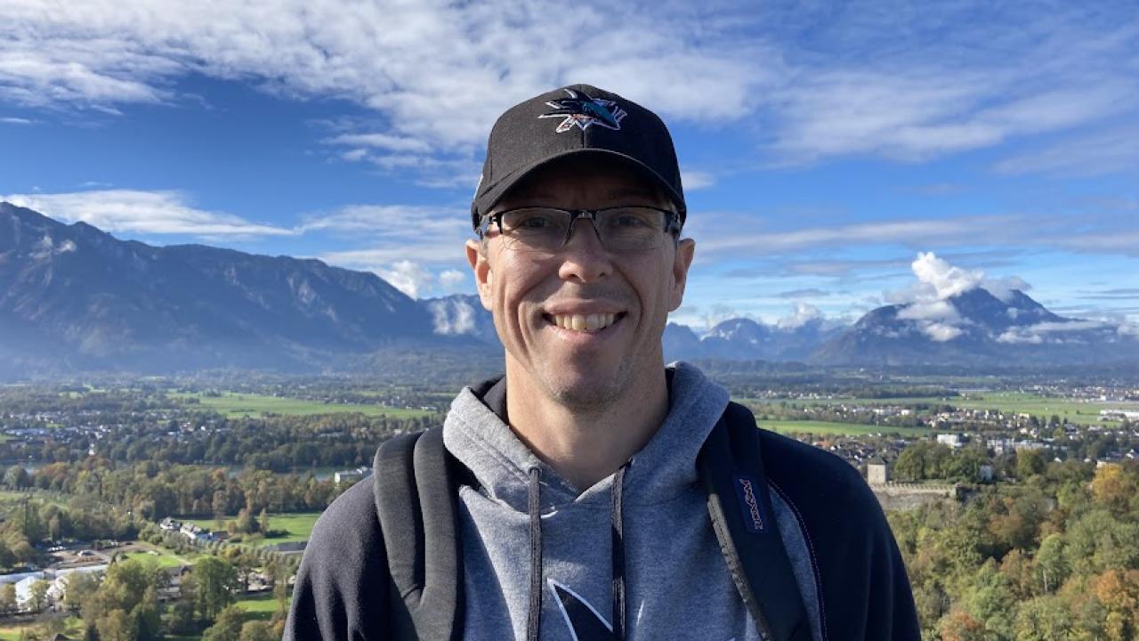 Scott smiles in a black San Jose Sharks baseball cap and sweatshirt in front of a scenic view with mountains in the background.