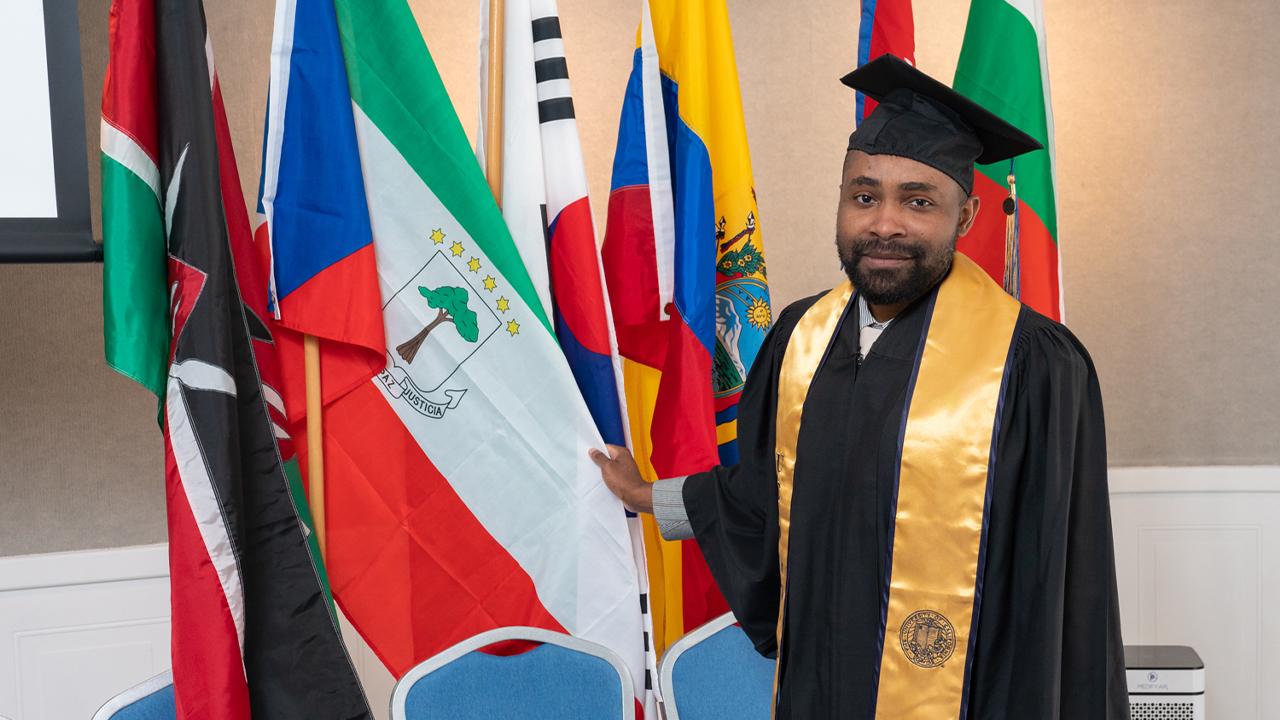 Edu stands in a black cap and gown next to a row of country flags. He holds his country flag out and looks at the camera.