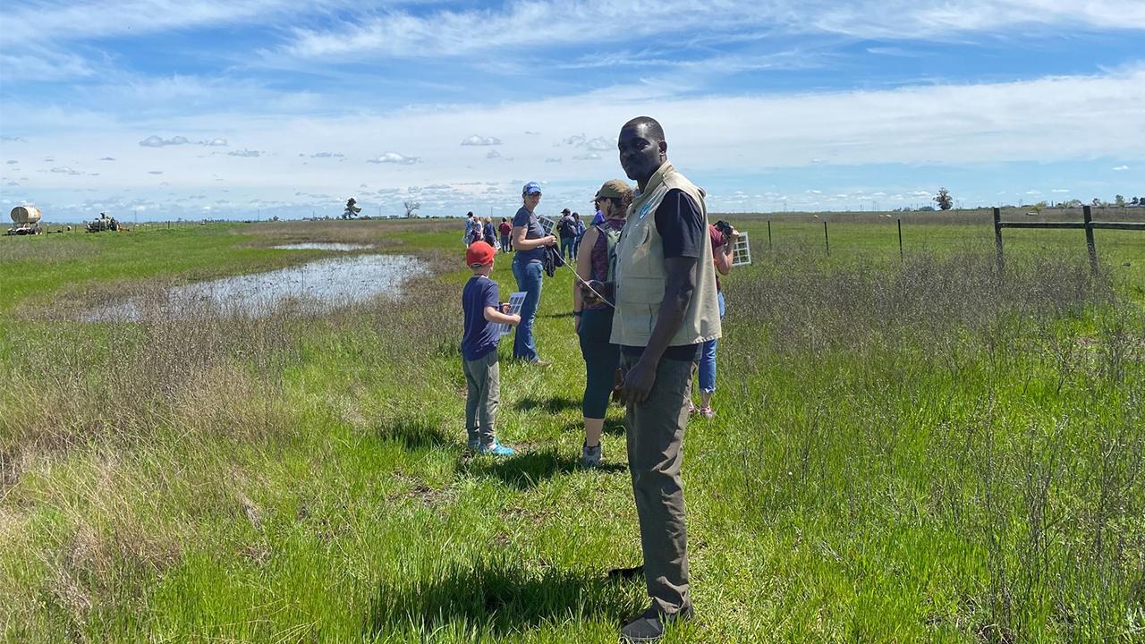 Silas stands in the middle of grassy wetlands on a sunny day with a group of about a dozen people in the distance behind him. He wears a khaki work vest, and his body is half turned and he looks over his shoulder at the camera. The blue sky above is streaked with clouds overhead with some cumulus clouds in the distance. 