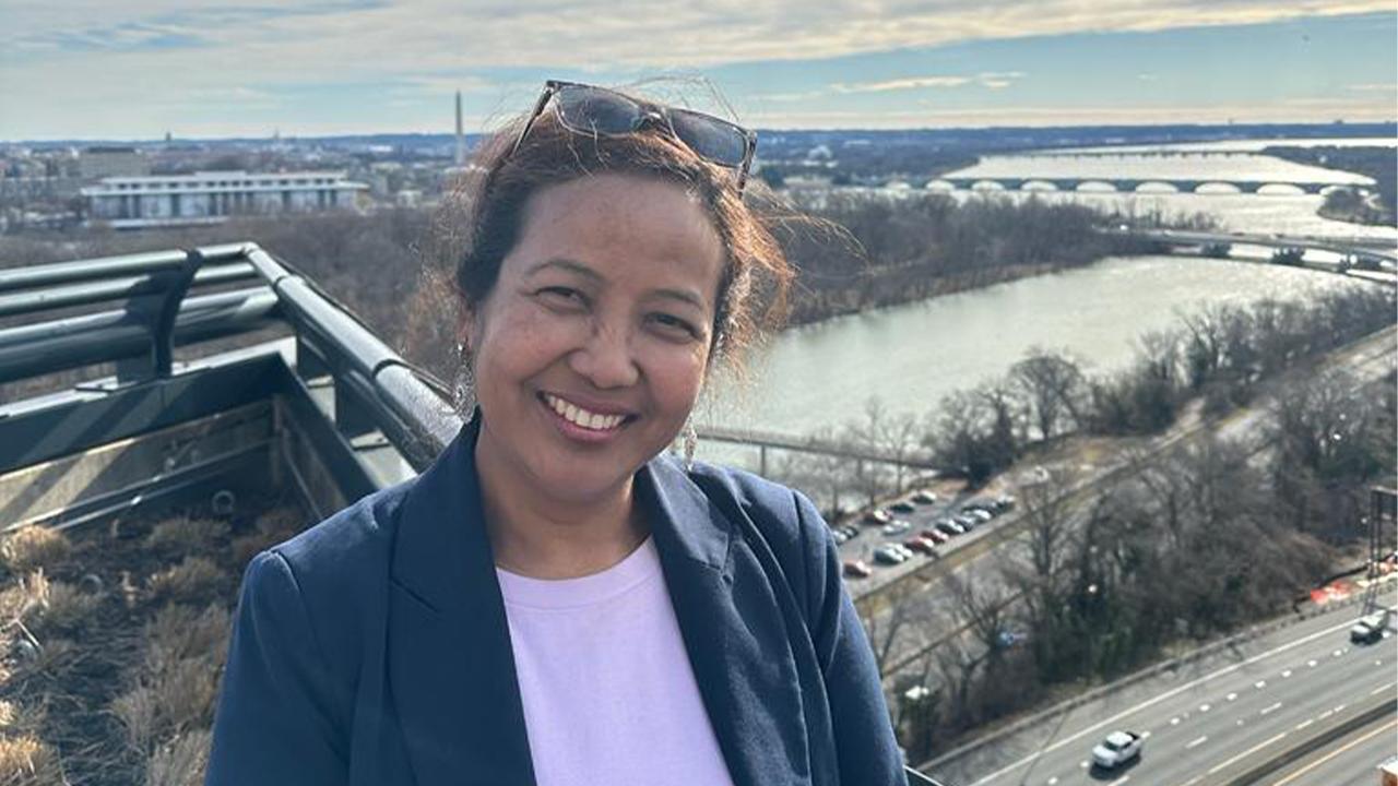 Sushila wears a white top under a dark blue blazer and smiles at the camera, Downtown Washington D.C. extends below and behind her with the Washington Monument off in the distance over her right shoulder.