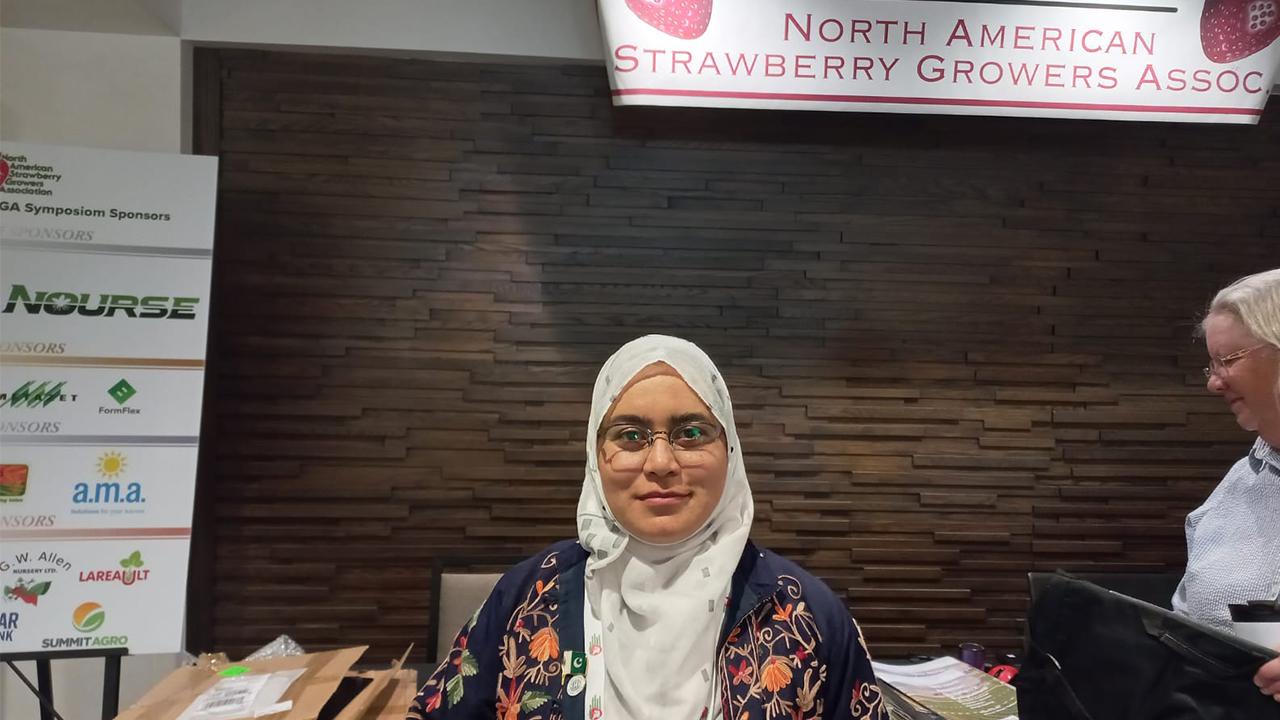 Nadia wears a hajib and stand in front of a dark wooden wall under a sign that reads "North American Strawberry Growers Association."