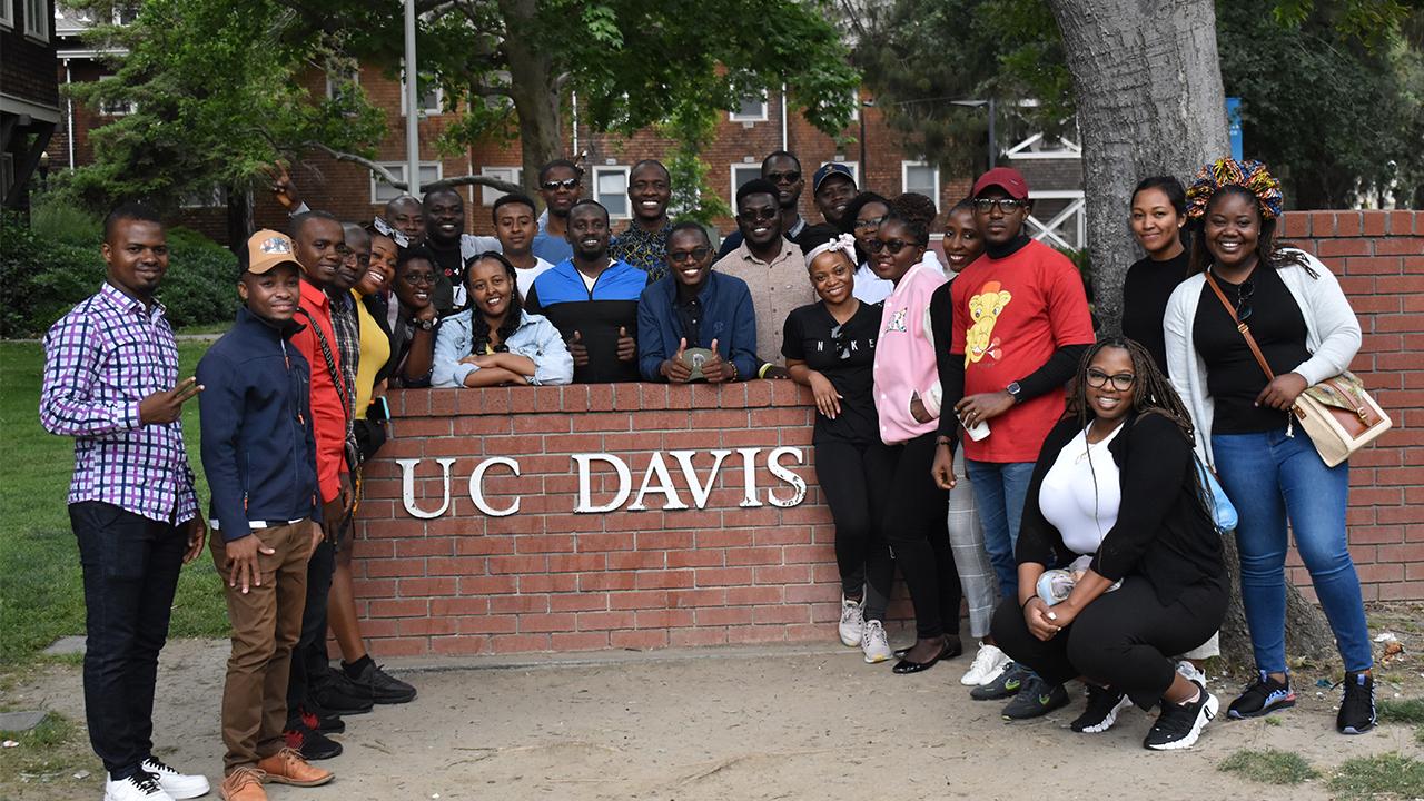 The group of 25 Fellows gathers around a short brick wall with a white UC Davis sign on it, surrounded by trees in the background.