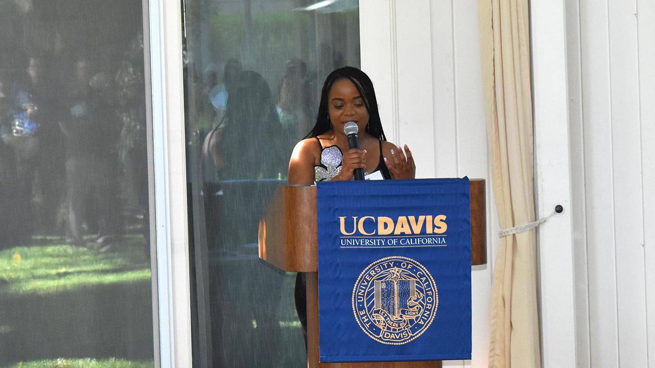Faith stands behind a wood podium with a blue drape that has the UC Davis seal and workmark in gold. She speaks into the microphone she is holding as she glances at her notes. 