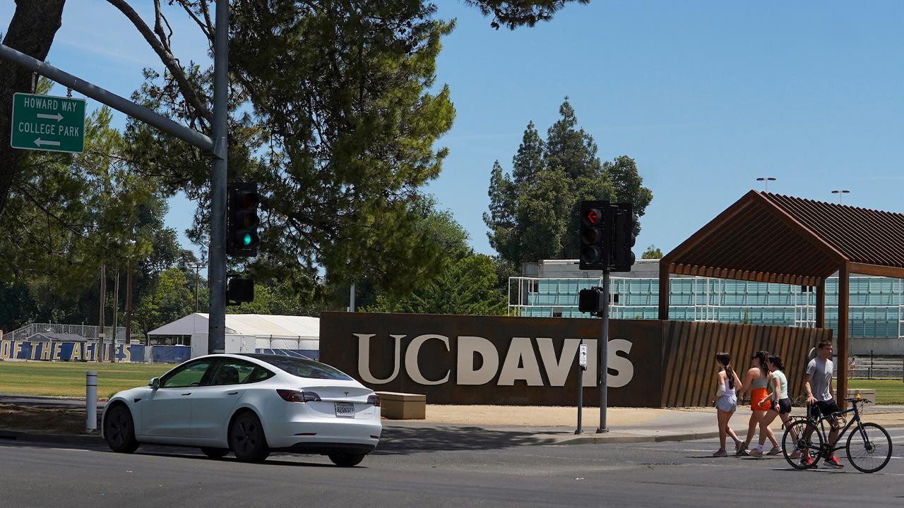 The UC Davis sign on a concrete wall on the corner of College Park and Howard Way at one of the entrances to the UC Davis campus on a sunny day. Three students on foot cross the street in the crosswalk toward the sign and another student walks a bike across the crosswalk away from the sign. A white Tesla crosses the street heading east past the sign.