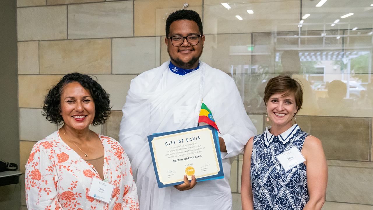 Gloria (left) wears a flowy floral top in white with pink flowers. Abnet (middle) wears a white tunic and pants with a sheer white overlay draped around his neck and over one shoulder. His collar is blue with large white clover patterns and he wears glasses. He holds a certificate proclaiming him an honorary citizen of Davis. Shelly (right) wears a sleeveless blue and white floral dress with a white scalloped collar with blue stripes. The trio stands inside in front of a wall of polished stone tiles.