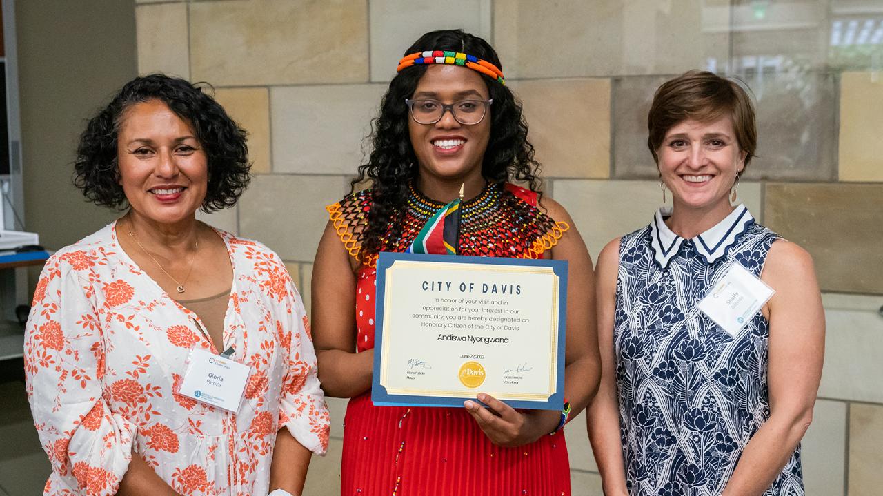 Gloria wears a floral top in white with pink flowers. Andiswa stands next to her in a red top with white polka dots, a red pleated skirt, a woven collar in black and orange, black glasses and a colorful headband. She holds a small South Africa flag and a certificate proclaiming her an honorary citizen of Davis. Next to Andiswa, Shelly wears a sleeveless blue and white floral dress with a white scalloped collar with blue stripes. The trio stands inside in front of a wall of polished stone tiles.