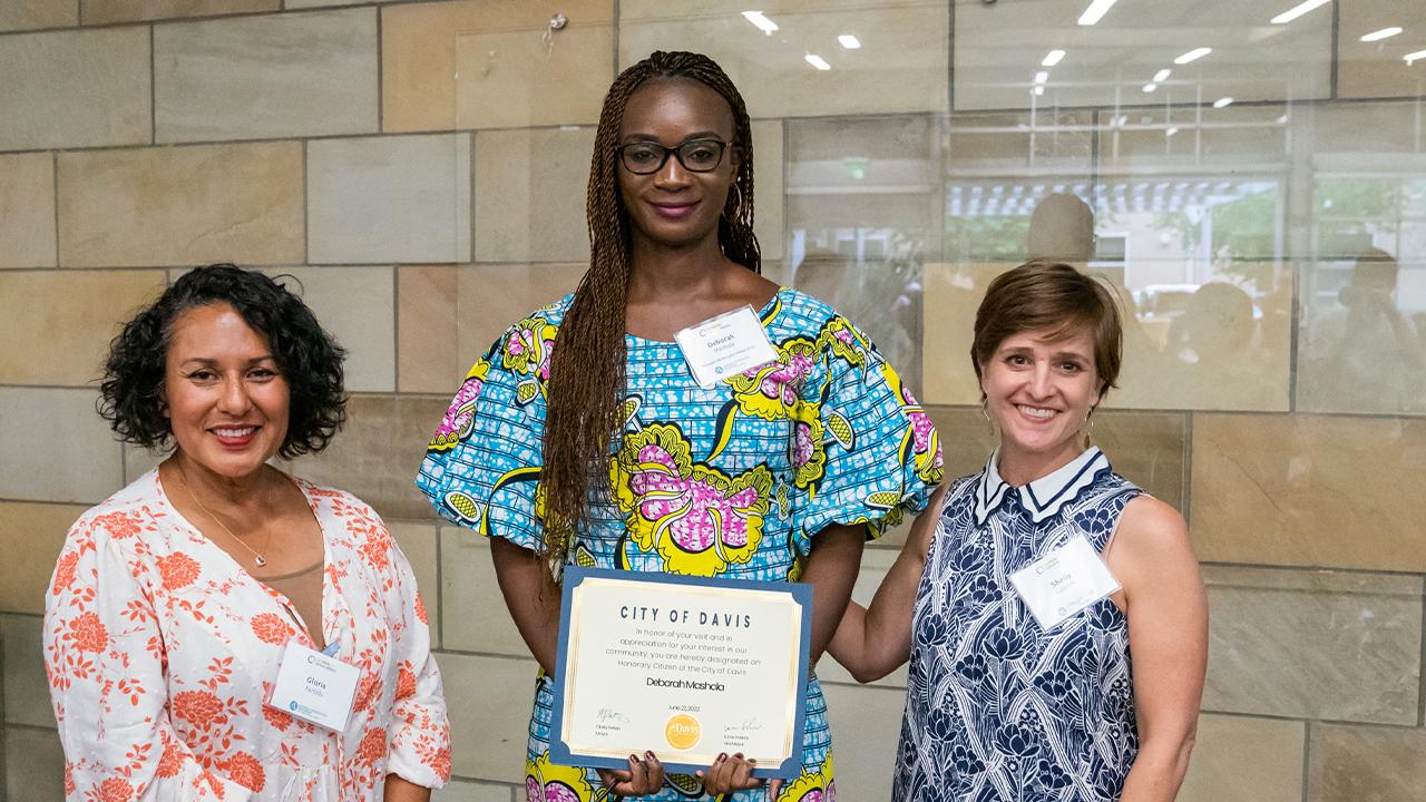 Gloria wears a flowy floral top in white with pink flowers. Deborah stands next to her in a traditional bright blue dress with large floral designs in yellow and dark pink and voluminous sleeves. She wears black rectangular glasses. She holds a certificate proclaiming her an honorary citizen of Davis. Next to Deborah, Shelly wears a sleeveless blue and white floral dress with a white scalloped collar with blue stripes. The trio stands inside in front of a wall of polished stone tiles.