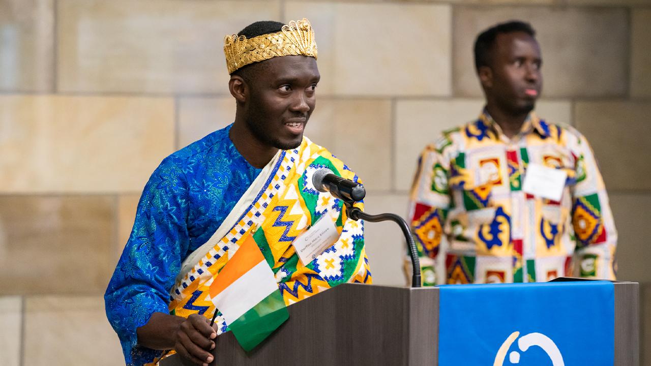 Marc wears a woven crown and bright blue tunic with a subtle floral pattern under a drape with tribal patterns in yellow, blue, green and white. He stands at a podium and holds a small Cote d'Ivoire flag.