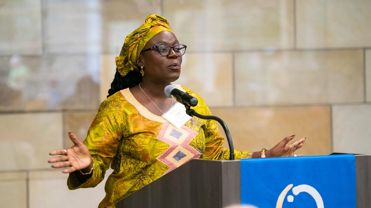 Kaustella wears a traditional Liberian headwrap and dress in yellow with and embellished patterned in gray and red. Down the front of the dress are diamond shapes in red, yellow and black. She wears metal framed glasses. She stands at a podium and is speaking. Her arms are spread wide as she gestures. 