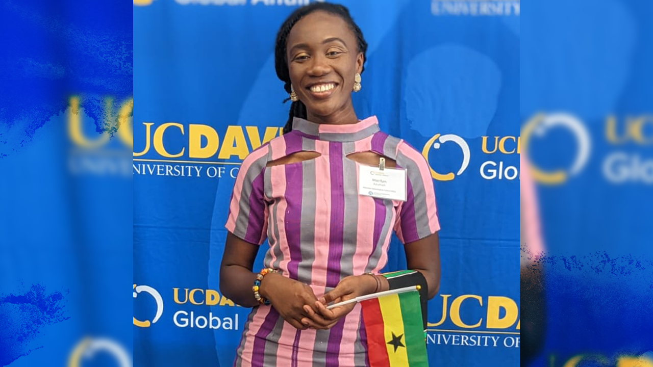 Marilyn wears a high-collar dress with thick vertical strips of light pink, magenta and gray. There are cut-outs over her collar bones. Her hands are clasped in front of her and she wears a colorful beaded bracelet on her right arm and holds in that hand a small Ghana flag. She smiles as she stands in front of a bright blue step-and-repeat board with yellow and white UC Davis and UC Davis Global Affairs logos.