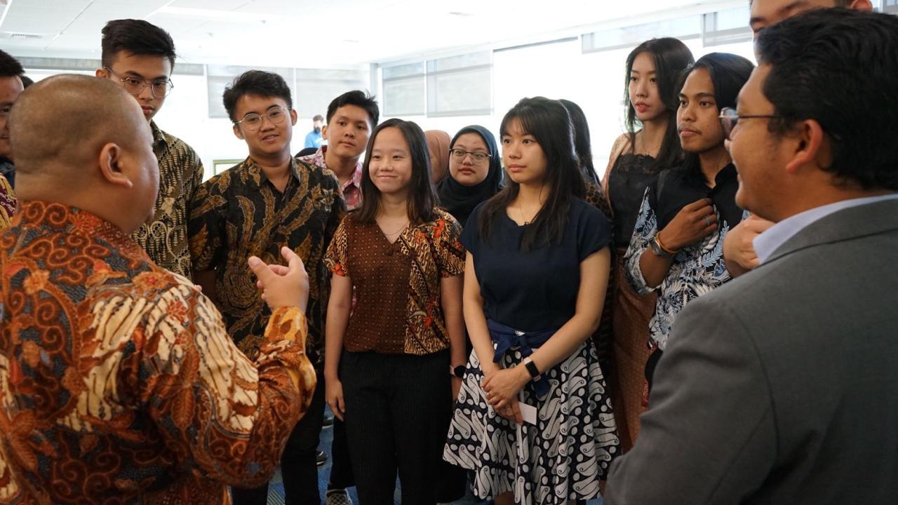 A group of a dozen male and female Asian students dressed in professional attire, many in patterns evocative of Indonesia culture, gather around a man in similar dress with his back to the camera at the left. The students, some smiling, are listening as the man gestures in an inclusive and welcoming way.